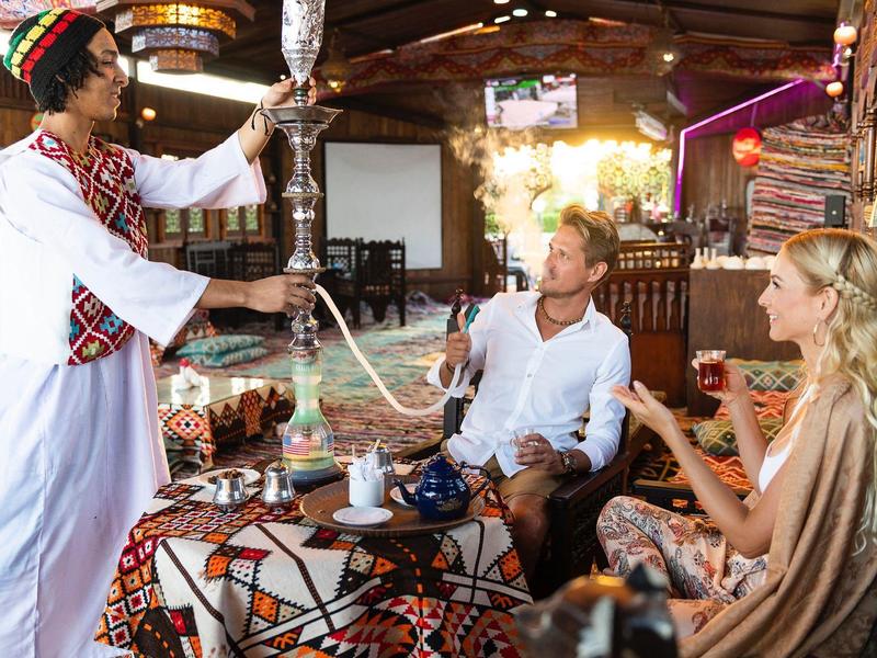 Ein Mann in traditioneller Kleidung bedient zwei Gäste mit einer Shisha in einem farbenfrohen, gemütlichen Raum.