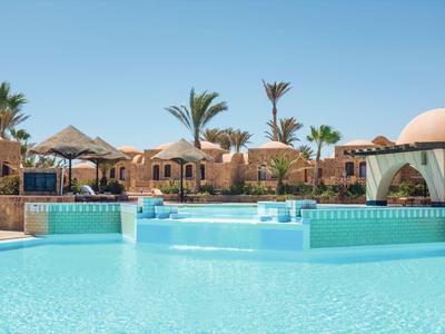 Grande piscine claire devant un hôtel avec des palmiers et des parasols au soleil.