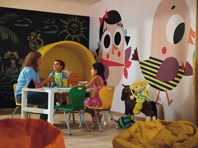 Niños sentados alrededor de una mesa en una habitación colorida y amigable para niños con murales y pufs.
