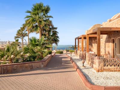 Un chemin pavé mène à un bâtiment aux murs en pierre naturelle, avec des palmiers et vue sur la mer.