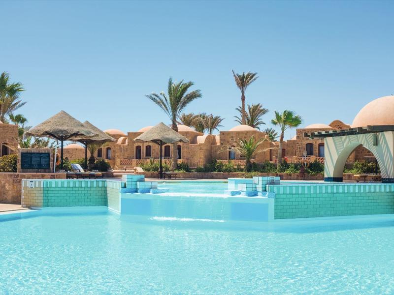 Gran piscina de hotel con agua azul clara, rodeada de palmeras y edificios beige bajo un cielo soleado.