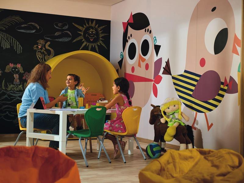 Niños sentados alrededor de una mesa en una habitación colorida y amigable para niños con murales y pufs.