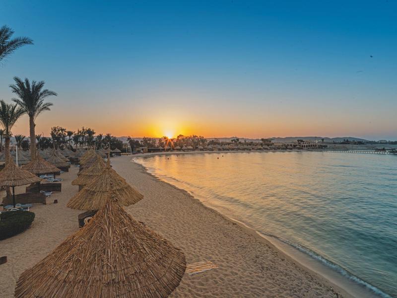 Playa con techos de paja, palmeras y agua tranquila al atardecer.