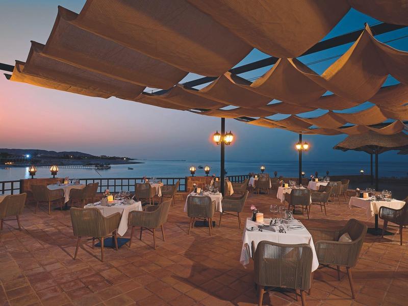 Restaurante al aire libre con mesas preparadas bajo toldos al atardecer junto al agua.