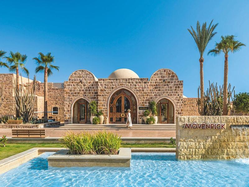 Hotel con fachada de piedra, varios arcos, palmeras y una piscina azul en primer plano.