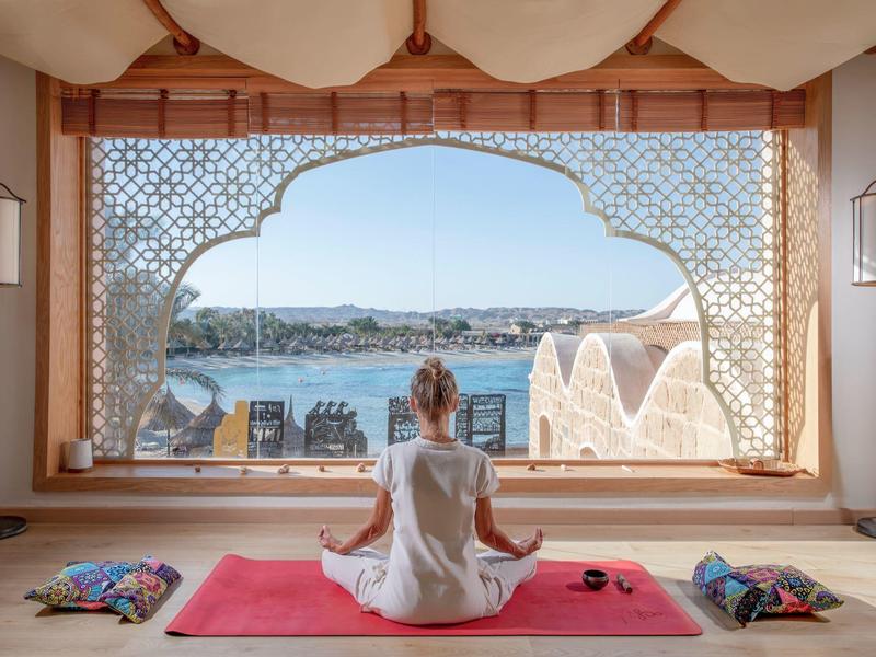 Mujer medita en esterilla con vista a un oasis y agua bajo un marco de ventana decorativo.