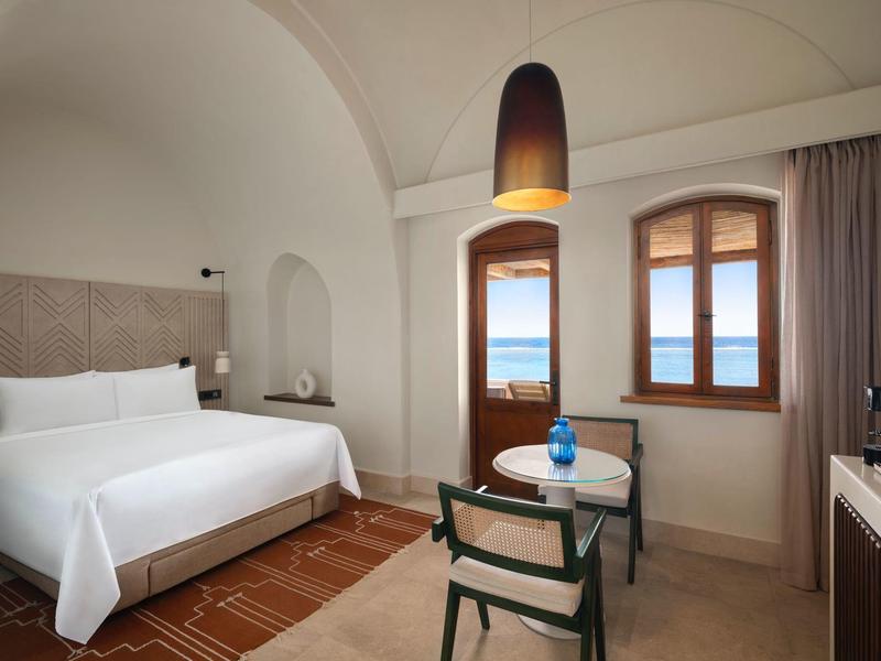 Habitación de hotel luminoso con cama doble, dos ventanas y área de asiento con mesa y sillas.