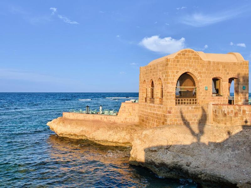 Edificio histórico de piedra en una península, rodeado por el mar azul y cielo despejado.