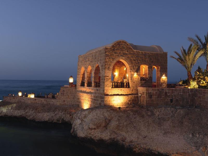 Casa de piedra iluminada en costa rocosa con palmeras al anochecer junto al mar.