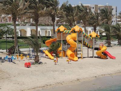 Area giochi colorata sulla spiaggia con palme e edifici sullo sfondo.