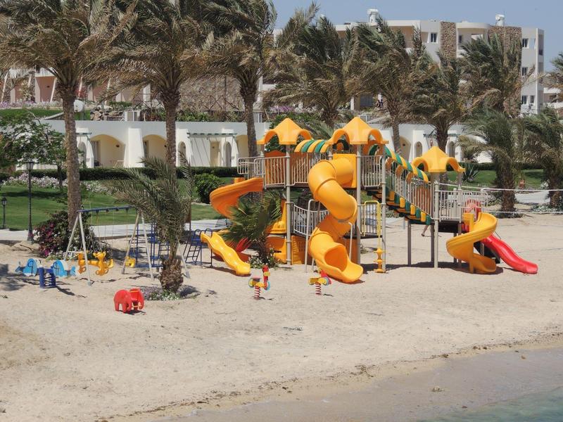 Area giochi colorata sulla spiaggia con palme e edifici sullo sfondo.