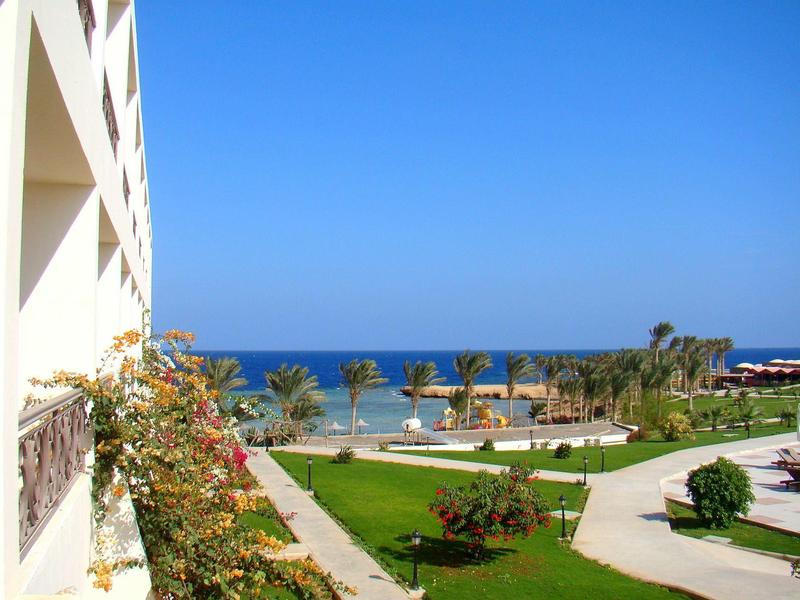 Vista su un giardino dell'hotel con sentieri verdi, palme e mare blu sullo sfondo.