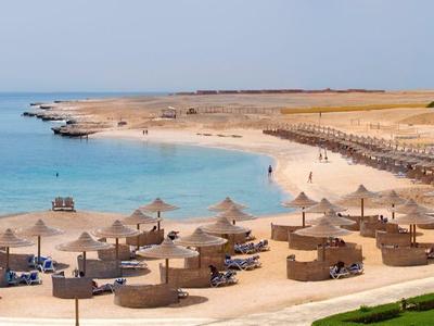 Strand mit Sand, Holzliegen, türkisfarbenem Meer und klarem Himmel, dazu grüne Wiese im Vordergrund.