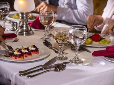 Weiß gedeckter Tisch mit Teller, Gläsern und Dessert, umgeben von Menschen beim Essen.