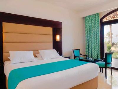 Helles Schlafzimmer mit großem Bett, türkisfarbenem Läufer, zwei Sesseln und grün gemusterten Vorhängen.