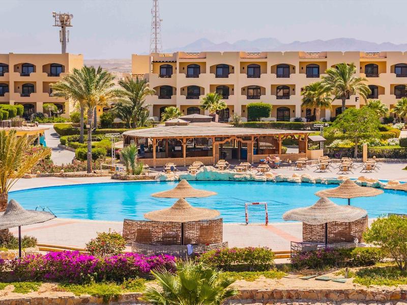 Duży okrągły basen z parasolami przed hotelem i palmami.