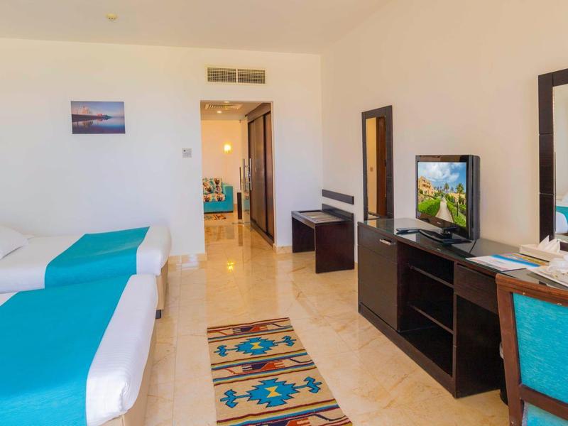 Jasny pokój hotelowy z dwoma łóżkami, biurkiem, telewizorem i turkusowymi akcentami.