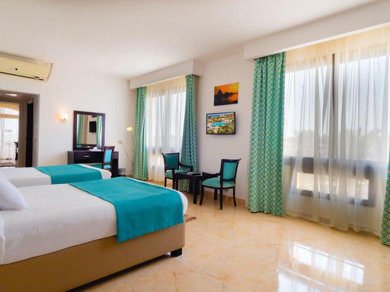 Przestronny pokój hotelowy z dwoma łóżkami podwójnymi, biurkiem i dużymi oknami z zielonymi zasłonami.