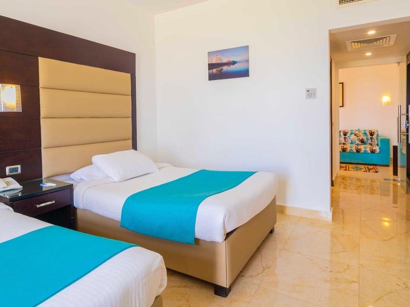 Pokój hotelowy z dwoma łóżkami pojedynczymi, niebieskimi akcentami i korytarzem do innego pokoju.