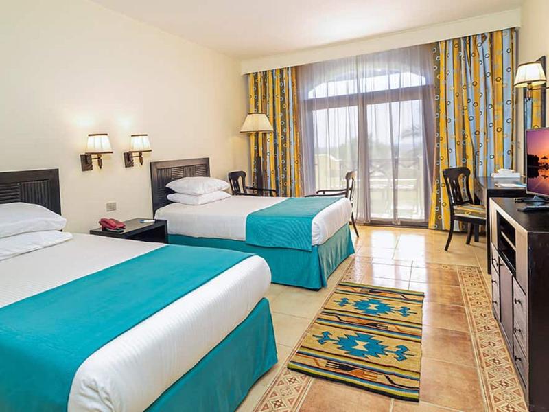 Jasny pokój hotelowy z dwoma łóżkami, dywanikiem i balkonem z widokiem na morze.