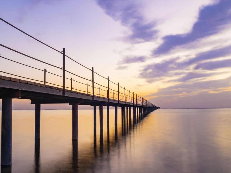 Długi pomost nad spokojną wodą o zachodzie słońca przy fioletowym niebie.