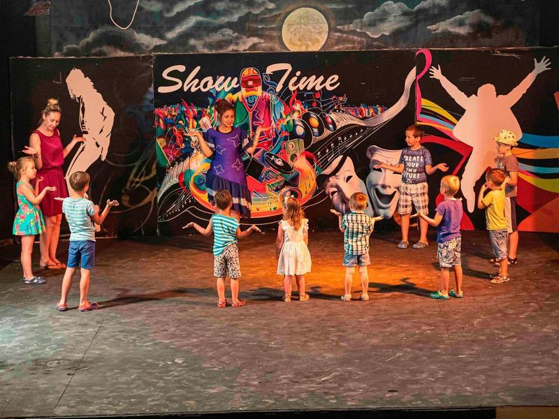 Grupa dzieci i dorosłych przed kolorową ścianą z napisem "Show Time" w sali.