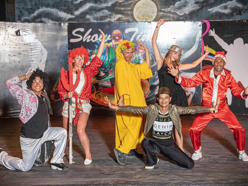 Sześć młodych osób w kolorowych strojach radośnie pozuje na scenie tanecznej.