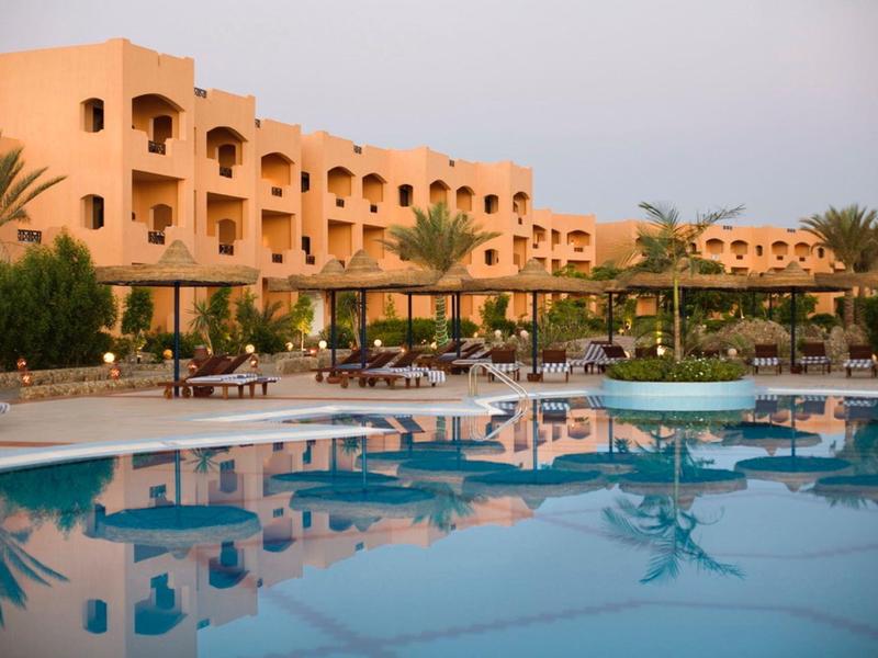 Hotel z basenem otoczonym palmami i leżakami pod pergolami o zmierzchu.