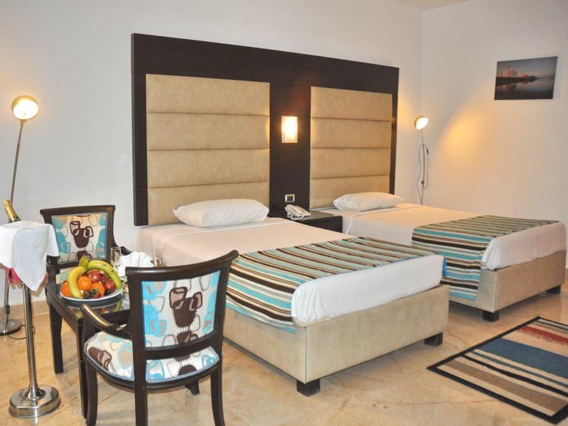 Pokój hotelowy z dwoma pojedynczymi łóżkami, krzesłami, koszem owoców i prostokątnymi dywanami.