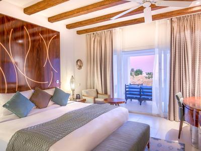 Bright hotel room with double bed, wooden beams, curtains, and balcony overlooking the sea.