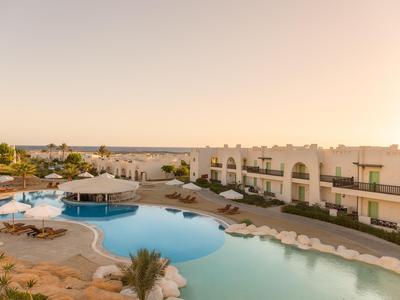View of a resort pool area with adjacent hotel rooms and the sea in the background at sunset.