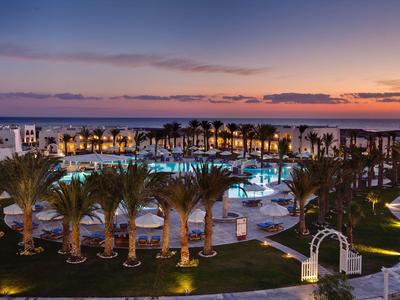 Luxury hotel complex with illuminated pool and palm trees at sunset by the sea