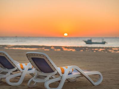 Two white lounge chairs on the beach overlooking sunset and a boat on the sea.