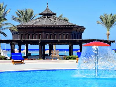 Une piscine avec un jeu d'eau devant une terrasse couverte et des palmiers près de la mer.