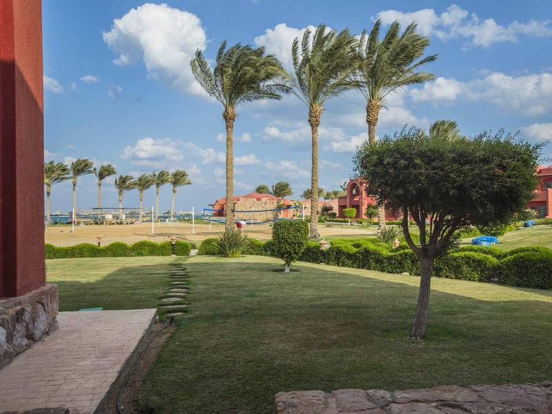 Vue sur une pelouse bien entretenue avec des palmiers et des bâtiments en arrière-plan sous un ciel bleu.