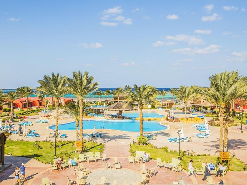 Complex hôtelier avec une grande piscine, des parasols et des palmiers sous un ciel clair.