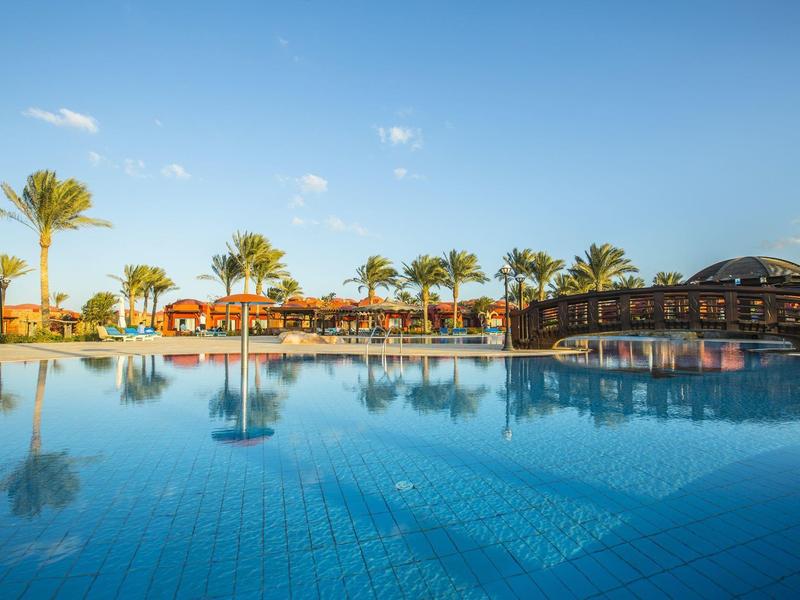 Une grande piscine avec de l'eau claire, entourée de palmiers et de bâtiments rouges sous un ciel bleu.