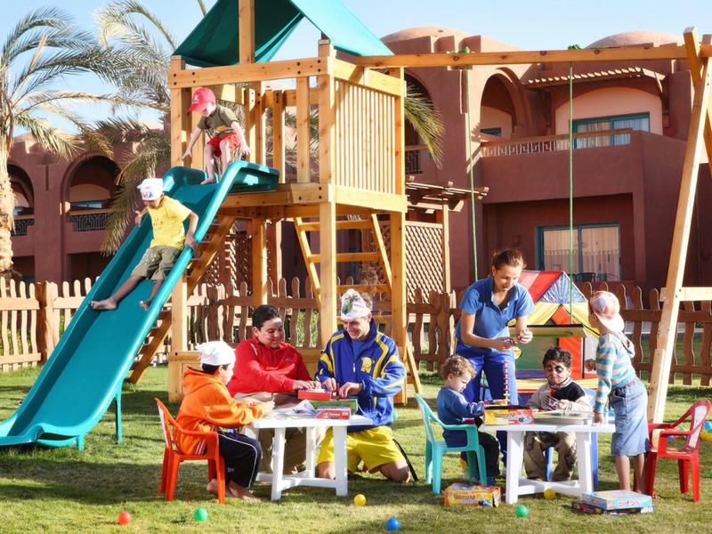 Des enfants jouent sur une aire de jeux avec toboggan et structure d'escalade dans un jardin d'hôtel.