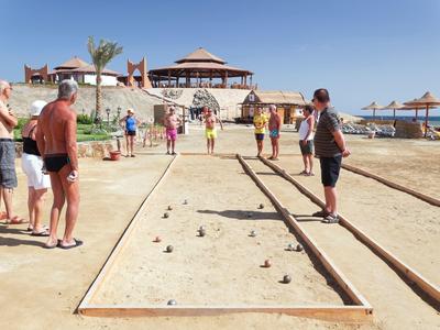 Menschen spielen Boccia auf Sandplatz am Strand mit Sonnenschirmen und Restaurant im Hintergrund.