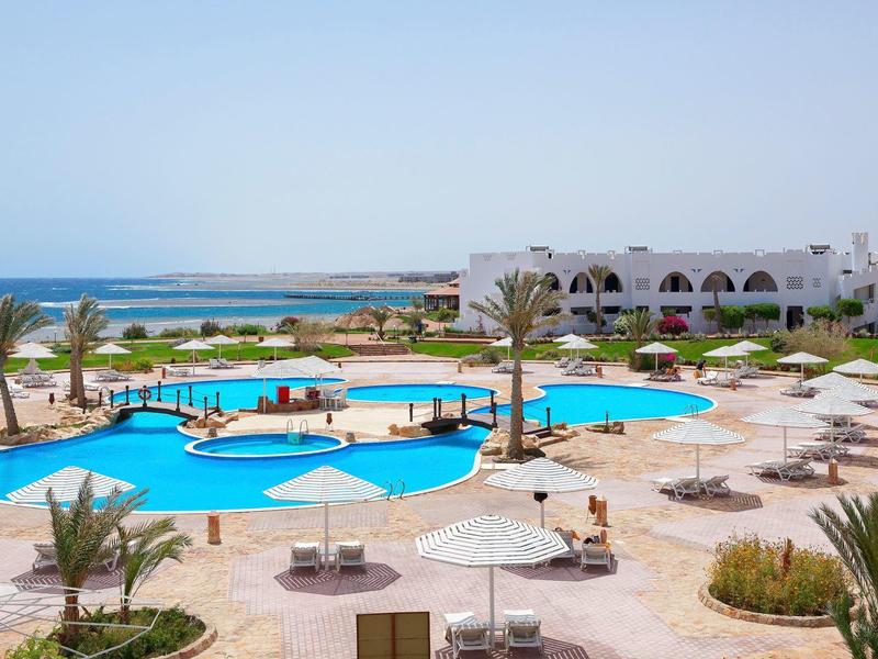 Piscina all'aperto con lettini, palme e edificio bianco vicino al mare.