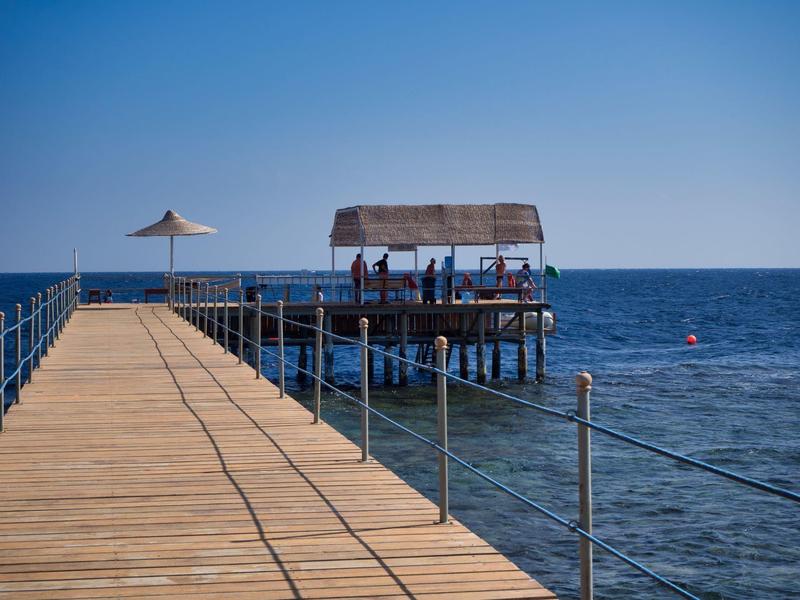 Molo s deštníkem a malým pavilonem nad průzračným mořem pod modrou oblohou.