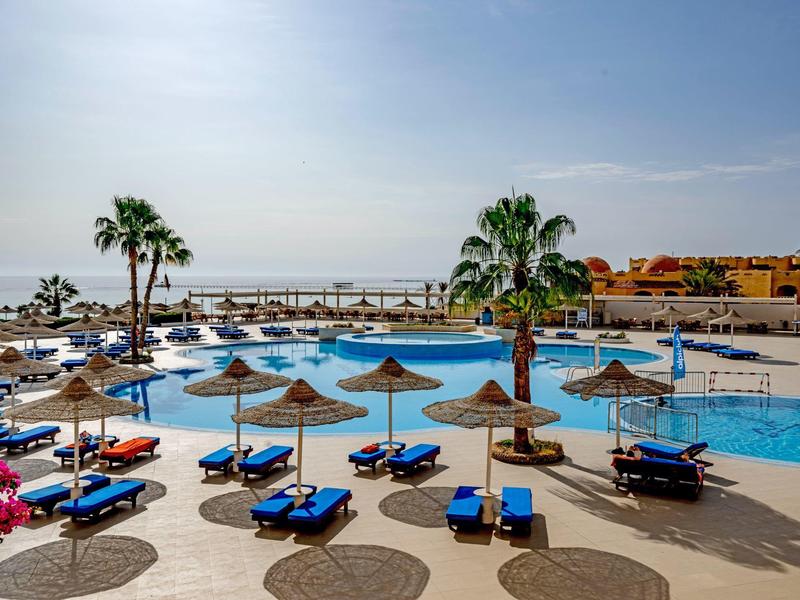 Bazénová oblast s modrými lehátky a slunečníky u moře pod jasnou oblohou.