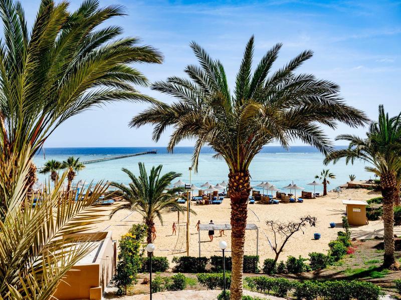 Písčitá pláž s palmami a výhledem na klidné modré moře pod jasnou oblohou.