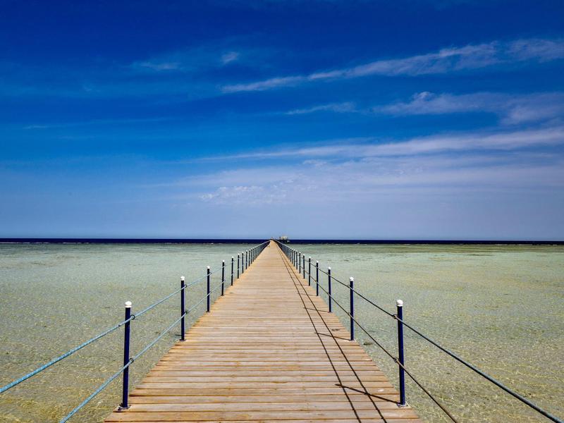 Dlouhý dřevěný můstek vede přes mělkou vodu pod jasnou modrou oblohou.