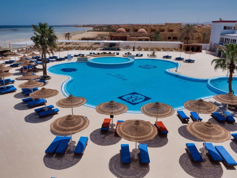 Velký hotelový bazén s modrými lehátky a slaměnými slunečníky na slunné pláži.