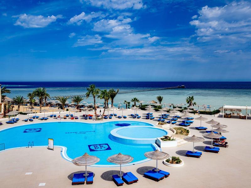 Velká oblast bazénu s modrou vodou, lehátky a výhledem na moře za jasné oblohy.