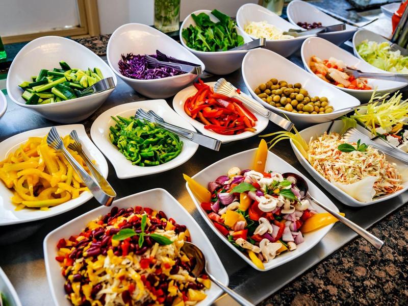 Barevný salátový bufet s různými nasekanými zeleninami a luštěninami v čtvercových miskách.