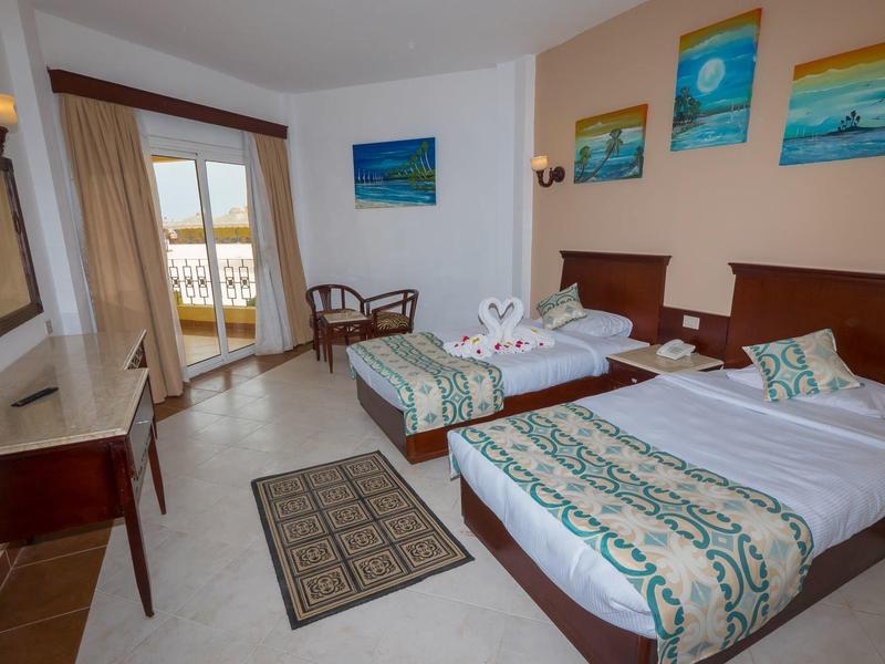 Hotelový pokoj se dvěma jednolůžky, balkonem a výhledem na moře, zdobený barevnými obrazy.