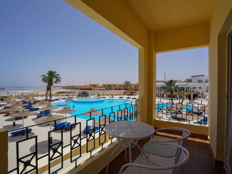 Balkon s stolem a židlemi s výhledem na bazén, lehátka a pláž za slunečného počasí.