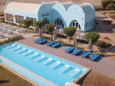 Piscina al aire libre con tumbonas y zona soleada junto a un edificio con fachadas azules.